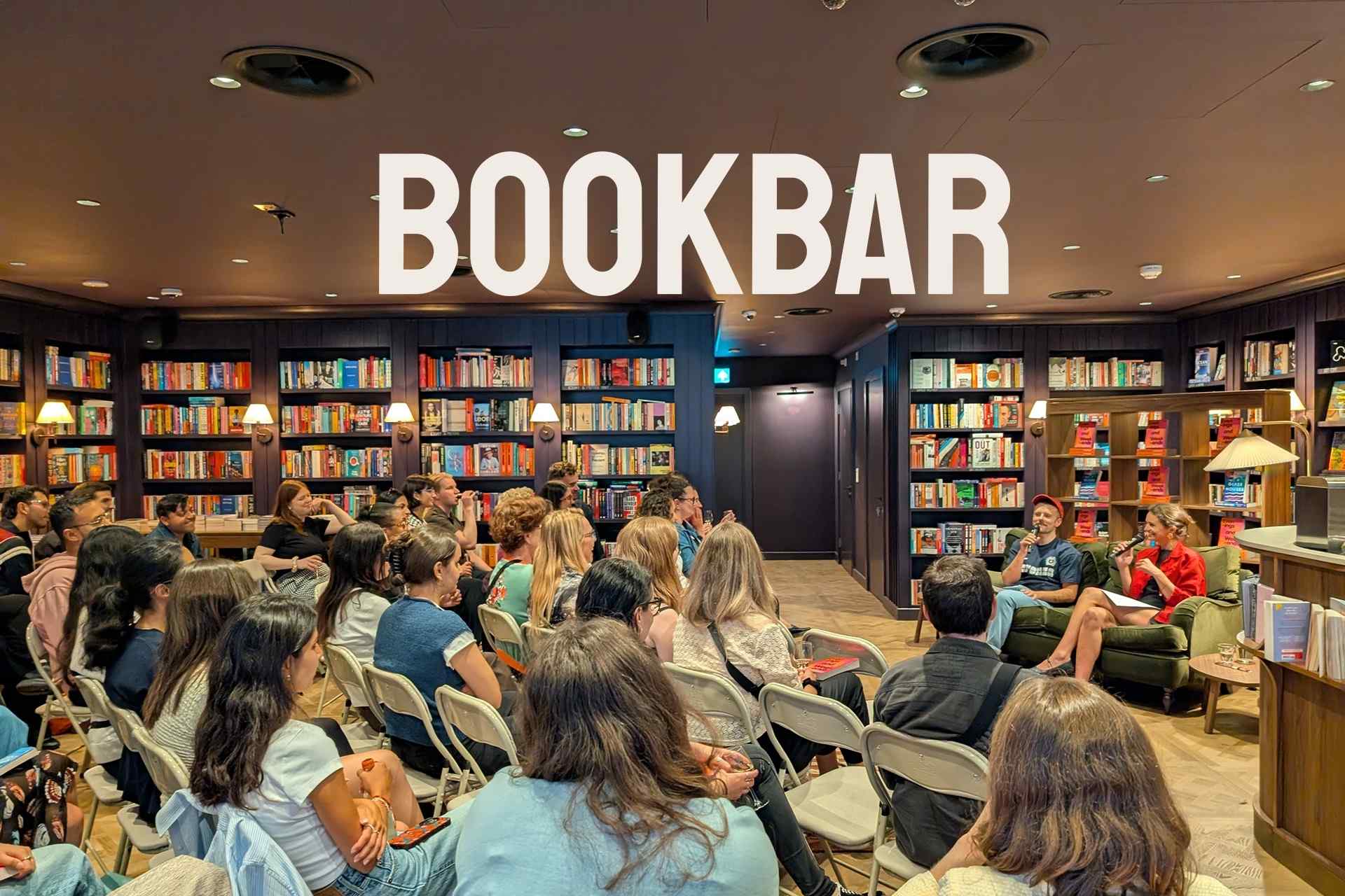 A crowded author event at Bookbar, an independent bookshop with floor-to-ceiling shelves of colorful books lining the walls. An audience of approximately 30 people sits in folding chairs facing two speakers seated on a green sofa, one holding a microphone. Warm wall sconces illuminate the dark wood-paneled interior.